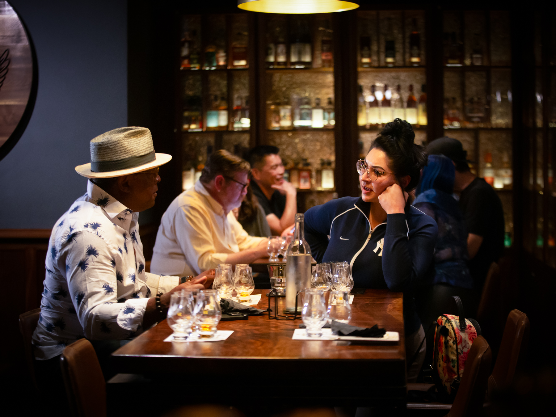 Customers sit around a table talking about the Whiskey they are tasting.  Whiskey bottles on shelves can be seen in the background